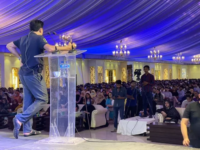 A speaker at a clear podium on a lit stage in front of a large audience